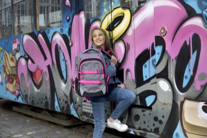 Mädchen steht mit Ihrem coolen neoxx Active Schulrucksack im Design Pink and Famous vor einem Zug, der mit Graffiti besprüht ist.